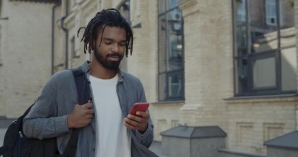 Crop view of afro american man with bag on shoulder typing message on smartphone screen and smiling. Bearded guy with dreadlocks using phonefor chatting while walking at city street. — Stock Video