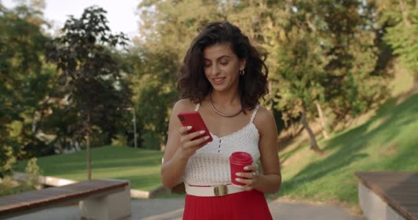 Jolie femme latino aux cheveux bruns ondulés utilisant un smartphone et portant une tasse de papier café. Femme naviguant sur Internet et écran tactile tout en marchant dans le parc de la ville. — Video