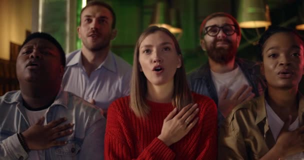 Grupo de amigos alegres cantando hino nacional enquanto sentados no pub. Diversas pessoas positivas dando alta cinco e olhando para a tela da TV enquanto se reúnem assistindo jogo de esportes. — Vídeo de Stock