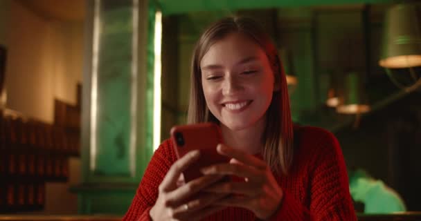 Close-up uitzicht op aantrekkelijke jonge vrouw glimlachen tijdens het scrollen en aanraken smartphone scherm. Gelukkige vrouwelijke persoon surfen op internet terwijl zitten aan tafel met bier en popcorn in de pub. — Stockvideo