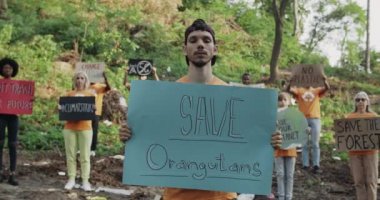 Başlık takan genç adam, çevrecilerin önünde dikilirken orangutanları kurtarıyor. Sancakları ve sloganları olan bir grup insan çöplerle dolu bir parkta kameraya bakıyor..