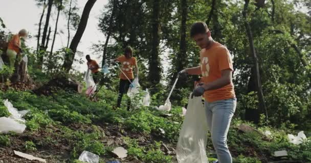 Young man using trash picker while collecting garbage in bin bag. Group of multiracial volunteers in t shirts with recycling symbol cleaning public park. Concept of cleanup and ecology. — Stock Video