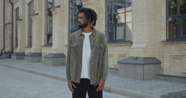 Vista de la cosecha de un joven con la barba girando la cabeza y mirando a la cámara. Retrato de un tipo afroamericano con rastas poniendo las manos en los bolsillos mientras está parado en la calle. Concepto de estilo de vida. — Vídeos de Stock