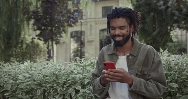 Νεαρός χαρούμενος τύπος με dreadlocks πληκτρολογώντας μήνυμα στην οθόνη του τηλεφώνου, ενώ στέκεται σε εξωτερικούς χώρους. Γενειοφόρος μικτή φυλή άνθρωπος σε casual ρούχα χρησιμοποιώντας smartphone και κουβέντα στα μέσα κοινωνικής δικτύωσης. — Αρχείο Βίντεο