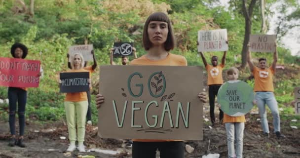 Mladá žena s kroužkem v nose, držící cedulku, je veganská, když stojí před ekologickými aktivisty. Skupina různorodých lidí s transparenty a slogany hledající kameru v parku plném odpadků. — Stock video