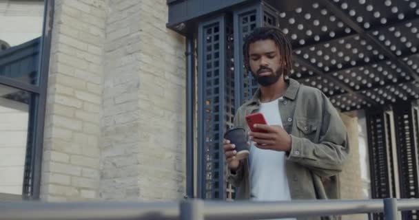 Νεαρός Αφροαμερικάνος με dreadlocks σε casual ρούχα που περιηγούνται στο διαδίκτυο και μεταφέρουν καφέ χάρτινο κύπελλο. Γενειοφόρος χαρούμενος τύπος χρησιμοποιώντας smartphone, ενώ το περπάτημα στο δρόμο της πόλης. — Αρχείο Βίντεο