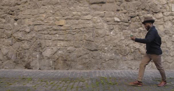 Hombre guapo en sombrero bailando y cantando mientras mira la pantalla del teléfono inteligente. Hombre afroamericano en ropa de moda escuchando música y moviéndose en rima mientras camina por la calle de la ciudad. — Vídeos de Stock