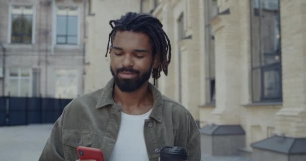 Crop view of young cheerful guy with dreadlocks holding paper cup and scrolling phone screen. Bearded handsome man smiling while using smartphone and walking at city street. — Stock Video