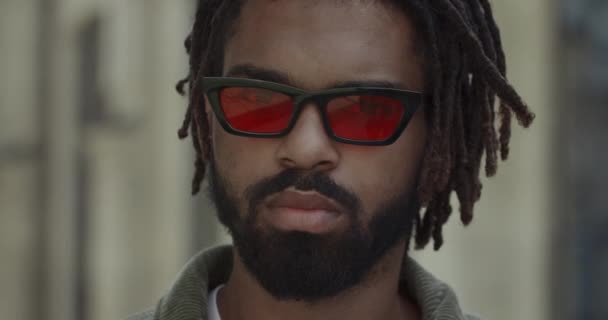 Close up view of man with beard and moustache turning head and looking to camera. Portrait of mixed race guy with dreadlocks wearing stylish glasses while standing at city street. — Stock Video