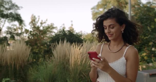 Vista Crop di bella donna scorrimento notizie di social media feed e accarezzare i capelli. Bella persona femminile bruna con capelli ondulati che toccano lo schermo e sorridono mentre camminano al parco della città. — Video Stock