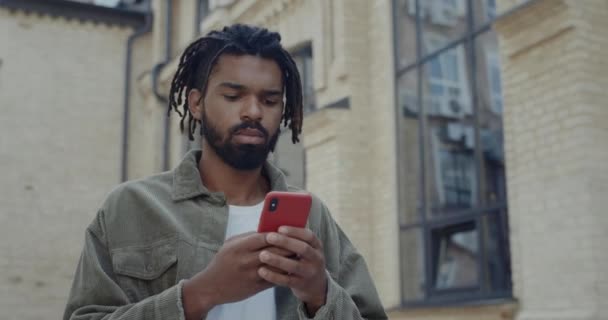 Crop view of young african american guy with dreadlocks typing message while chatting in social media. Bearded man using smartphone while walking at city street. Concept of communication. — Stock Video