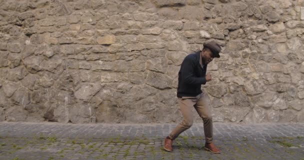 Överlycklig manlig dansare i hatt rör sig och sjunger på gamla stan gatan. Talangfull ung afro amerikan man i trendiga kläder har roligt medan stående city street. Begreppet lycka. — Stockvideo