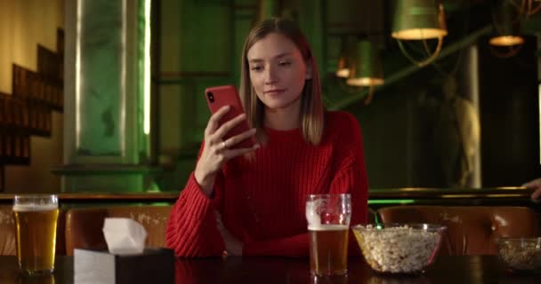 Hübsche junge Frau mit Smartphone am Tisch mit Bier und Popcorn darauf. Diverse Menschen lächeln und begleiten ihre gut gelaunte Freundin zum Sport in der Kneipe. — Stockvideo