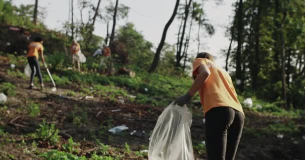 Vista trasera de una joven voluntaria usando recolector de basura mientras camina en el bosque. Grupo de eco activistas en las mismas camisetas recogiendo basura. Concepto de limpieza y ecología. — Vídeo de stock