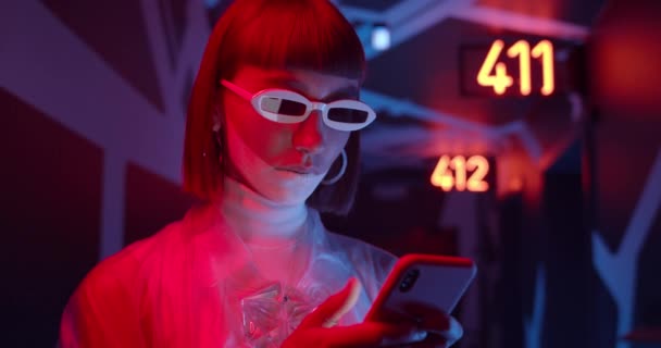 Portrait of female young person browsing internet whule standing at corridor with futuristic neon light. Trendy woman in party glasses looking at smartphone screen and than to camera. — Stock Video