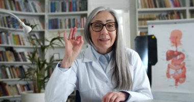 Gözlüklü olgun bir kadın doktor video konsültasyonu sırasında ekrana dokunmaktansa işaret veriyor ve el sallıyor. Beyaz önlüklü kadın, muayenehanede otururken konuşuyor..