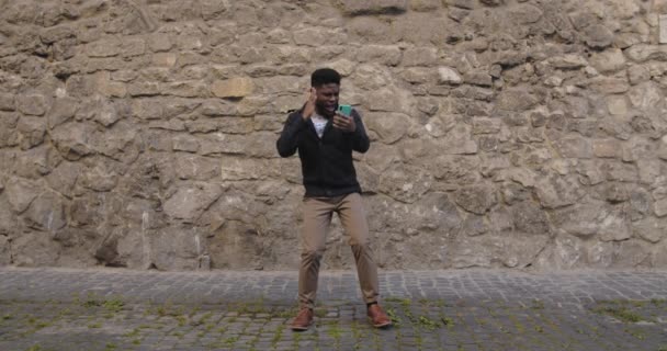 Telefona bakarken dans eden ve şarkı söyleyen olumlu bir erkek. Sakallı, şık giyinmiş, müzik dinleyen ve şehir caddesinde dururken kafiyeli hareket eden Afro-Amerikan bir adam.. — Stok video