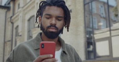 Elinde kahve fincanı olan ve sosyal medyada sohbet eden Afrikalı Amerikalı genç bir adamı yakından çek. Sakallı adam akıllı telefon kullanıyor ve sokakta yürürken gülümsüyor..