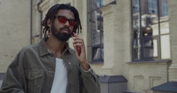 Young guy with dreadlocks dailing number on phone screen and smiling outdoors. Bearded man in stylish red glasses talking on smartphone while walking at city street. — Stock Video