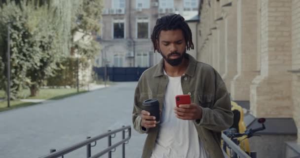 Beskär syn på ung afrikansk amerikansk man med dreadlocks surfa internet och bär kaffepapper kopp. Skäggig seriös kille som använder smartphone när du går på city street. — Stockvideo