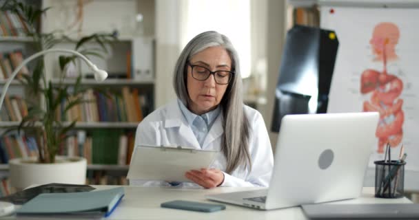 Kvinnliga äldre läkare talar och håller pappershållare samtidigt fylla i medicinska formuläret. Gammal dam i vit råtta lyssnar på patient och skriva information samtidigt som videosamtal. — Stockvideo