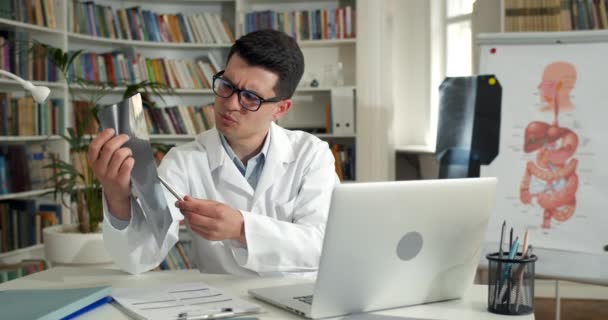 Médico varón sosteniendo la foto de rayos X del pecho mientras que tiene la consulta médica en línea en el oficina.Hombre joven en gafas y robo blanco usando el ordenador portátil mientras que se comunica con el paciente. — Vídeo de stock