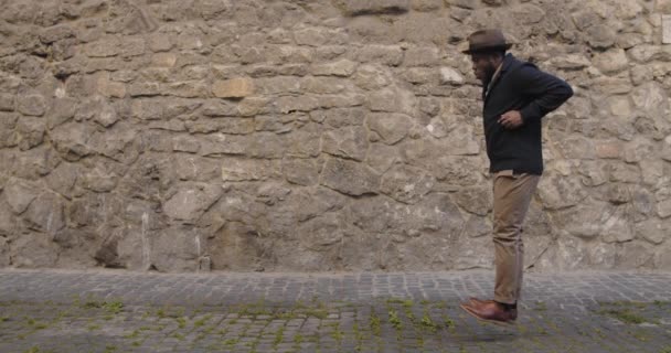 Hombre guapo en sombrero bailando y saltando mientras camina en la vieja calle de la ciudad. Chico afroamericano con ropa de moda moviéndose en rima. Concepto de buen humor, felicidad y diversión. — Vídeos de Stock