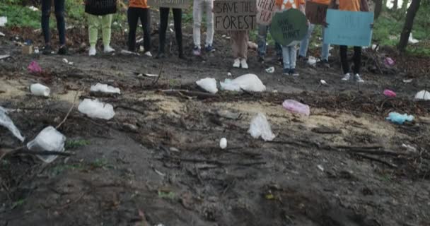 Group of diverse activists wearing t shirts with recycling symbol and holding carton placards with eco slogans. People looking to camera while standing in forest full of trash. — Stock Video