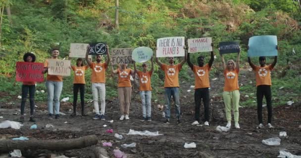 Különböző öko aktivisták viselnek pólók újrahasznosítás szimbólum bérbe karton plakátok öko szlogenekkel.Emberek csoportja, akik a kamera, miközben áll az erdőben tele szeméttel. — Stock videók