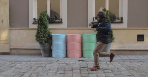 Männliche Person mit Hut tanzt und trinkt aus einer Pappkaffeetasse, während sie auf der Stadtstraße spaziert. Afro-amerikanischer stilvoller Mann, der sich in Reimen bewegt und kreist. Bunte Fässer im Hintergrund. — Stockvideo