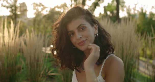 Vue rapprochée de la femme brune béatifique aux cheveux ondulés souriants et posant. Portrait de belle femme caressant les cheveux et regardant vers la caméra tout en étant assis dans le parc. Zoom avant. — Video