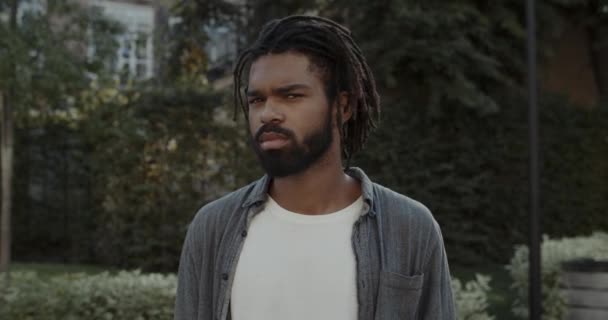 Crop view of handsome young man with beard turning head and looking to camera. Portrait of cheerful guy with dreadlocks smiling while standing at city street. Concept of lifestyle. — Stock Video