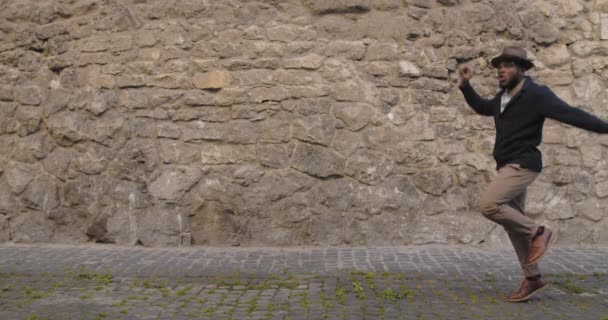 Handsome excited man in hat dancing, jumping and circling while walking at old city street. Afro american guy in fashionble clothes moving in rhytm. Concept of happiness and fun. — Stock Video