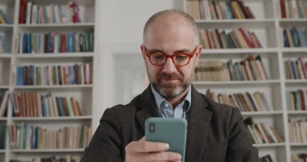 Közelkép a koncentrált öltönyös férfiról okostelefonnal. Portré idősebb szakállas férfi szemüveges olvasás hírek és megható képernyőn, miközben ül a szobában tele könyvek. — Stock videók
