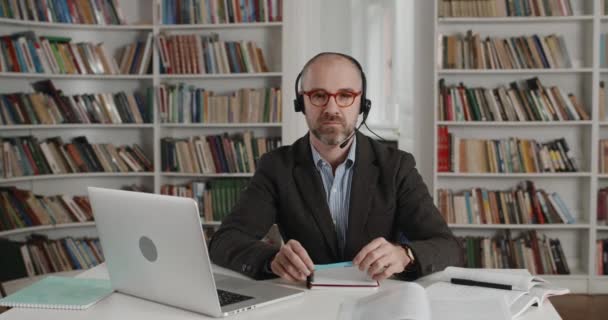 Portret van een vrolijke man in een headset met microfoon die naar de camera kijkt en glimlacht. Close-up weergave van volwassen bebaarde man in bril zitten aan een bureau in de buurt laptop in kamer vol boeken. — Stockvideo