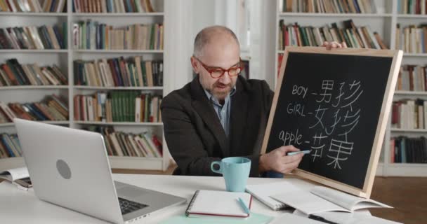 ノートパソコンを見ながらチョークと黒板を保持眼鏡で男の作物ビュー。男性教授はオンラインで言語を教える。教師の話の側面図。距離学習の概念. — ストック動画