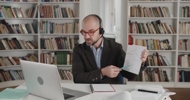 Egy szemüveges férfi, aki papírlapot tart háromszögekkel, miközben beszél és laptopot néz. Felnőtt férfi tanár fülhallgatóban, mikrofonos matektanítással online. A távoktatás fogalma. — Stock videók