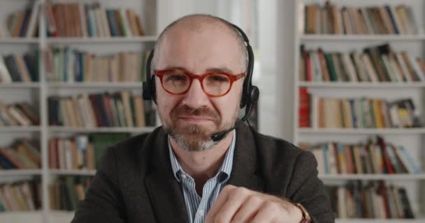 Close up of positive man in headset with mic looking to camera and smiling. Portrait of senior bearded male person in glasses sitting at desk near laptop in room full of books. — Stock Video