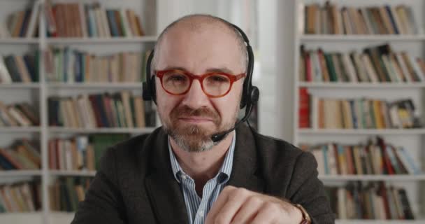 Primer plano del hombre positivo en auriculares con micrófono mirando a la cámara y sonriendo. Retrato de hombre barbudo mayor en gafas sentadas en el escritorio cerca de la computadora portátil en la habitación llena de libros. — Vídeos de Stock