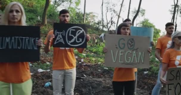 Des éco-activistes dans les mêmes t-shirts avec le symbole de recyclage debout dans le parc plein d'ordures. Groupe de personnes diverses tenant et soulevant des bannières en carton avec des slogans écologiques. — Video