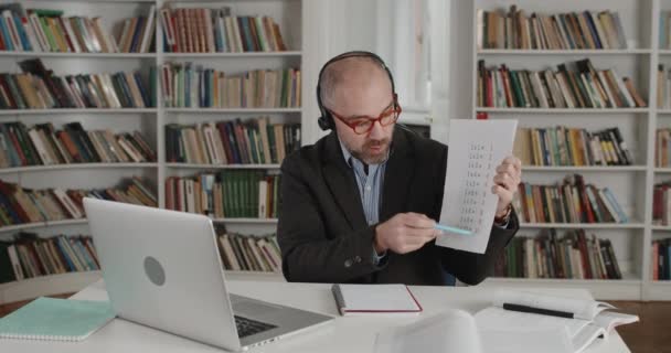 Crop view of man in glasses holding paper sheet with miltiplication table while talking and looking at laptop. Mature male teacher in headset with mic teaching math online. — Stock Video