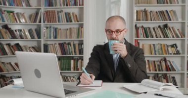 Takım elbiseli, gözlüklü, yetişkin bir profesörün kamera laptopu kullanırken dinlediği ekin görüntüsü. Adam kafa sallarken ve masa başında çalışırken not alıp içiyor. Çevrimiçi eğitim kavramı.