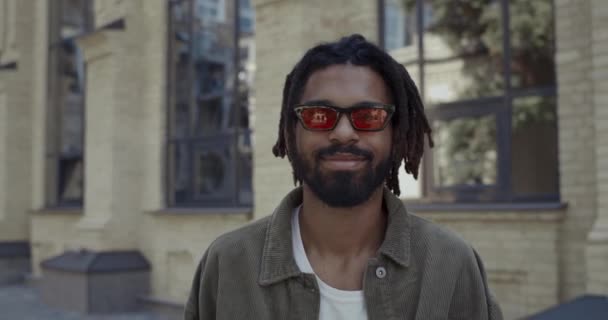 一个快乐的家伙,满面春风,面带笑容地看着外面的相机.头戴时髦红色眼镜的非洲裔美国人，在城市街道上散步时心情很好 — 图库视频影像