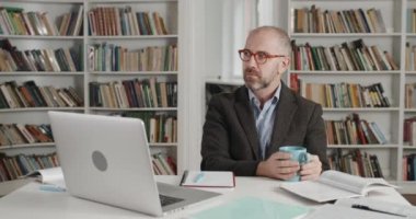 Takım elbiseli, gözlüklü, iletişim kuran ve kamera laptopu kullanan yetişkin erkek öğretmen görüntüsü. Masada çalışırken elinde bardak tutan bir adam. Çevrimiçi eğitim kavramı, öğrenme.