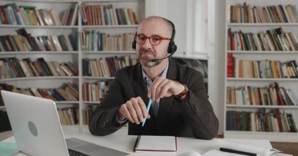 Retrato de hombre positivo en auriculares con micrófono girando la cabeza y mirando a la cámara. Vista cercana de hombre barbudo maduro en gafas sonriendo y sentado en el escritorio cerca de la computadora portátil. — Vídeo de stock