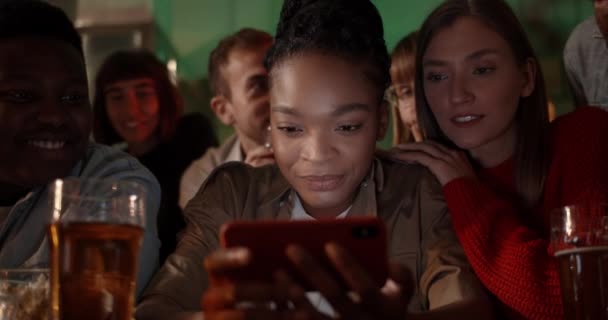 美国黑人女人和朋友坐在酒吧里看屏幕时，一边拿着电话一边笑的照片。多民族人群在智能手机上观看体育运动. — 图库视频影像