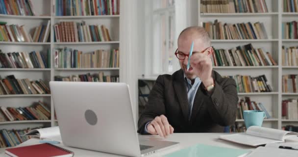 在使用笔记本电脑的时候，戴着眼镜、一边说话一边拿笔的快乐男人的照片。男教师穿着西服，坐在满是书的房间里，在网上上课。远程学习的概念. — 图库视频影像
