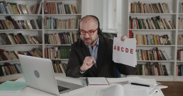 Vista de corte de homem sênior usando fone de ouvido com cartão de microfone com letras e usando câmera de laptop no escritório em casa. Pessoa masculina conversando e tendo aulas de vídeo. Conceito de educação online. — Vídeo de Stock