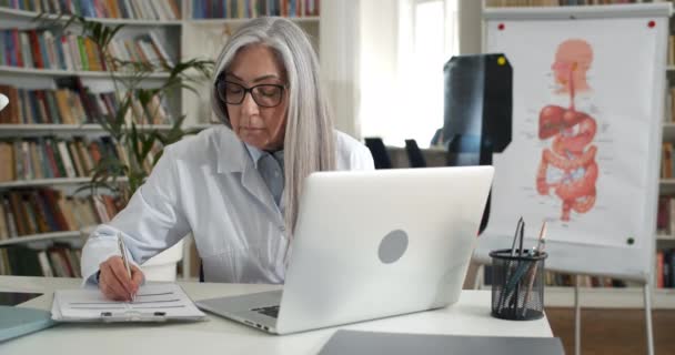 Mature concentrated woman in glasses and white gown typing and filling in medical documentation. Mature female doctor working with laptop in office. Concept of routine and work. — Stock Video