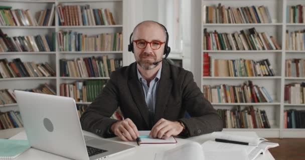 Retrato de profesor varón maduro positivo en auriculares con micrófono mirando a la cámara y sonriendo. Primer plano de hombre barbudo en gafas sentado en el escritorio cerca de la computadora portátil en la habitación llena de libros. — Vídeos de Stock